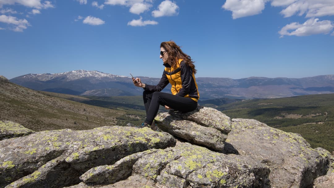 Kvnna i svarta kläder och gul väst som sitter på en sten med en mobiltelefon i handen. I bakgrunden stora berg. 