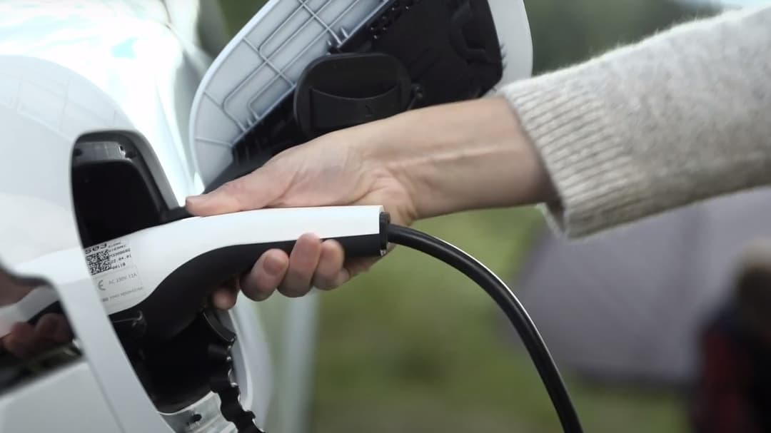A hand that plugs in a white electric car for charging. Nature and tent can be seen in the background.