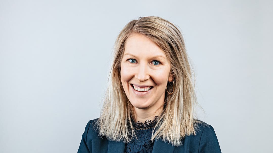 En kvinna i blå kavaj och med ett stort leende som sitter på en pall med händerna i knät