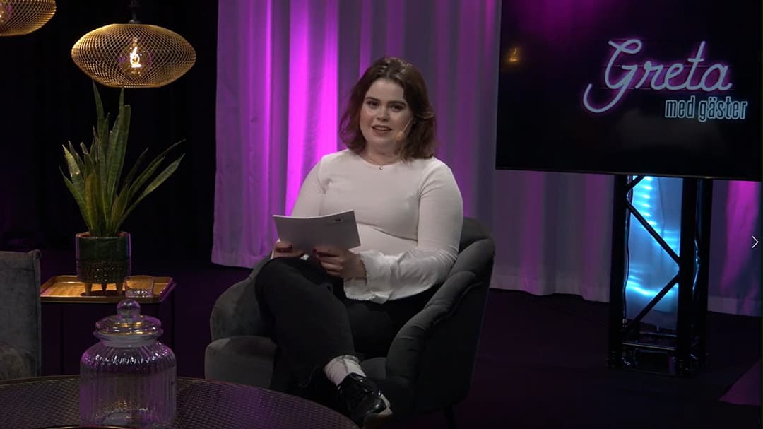 En kvinna sitter i en fåtölj i en studio, hon tittar in i kameran.
