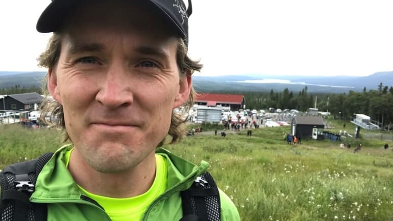 En man med keps och grön jacka med fjäll och polarull i bakgrunden 