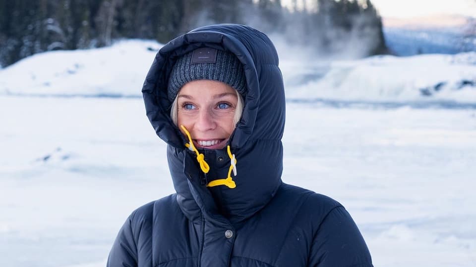 En kvinna i mörkblå täckjacka med huvan uppdragen och mössa står utomhus. Bakom henne finns ett snötäckt fält och längre bort granskog. Hon tittar åt sidan och ler. 