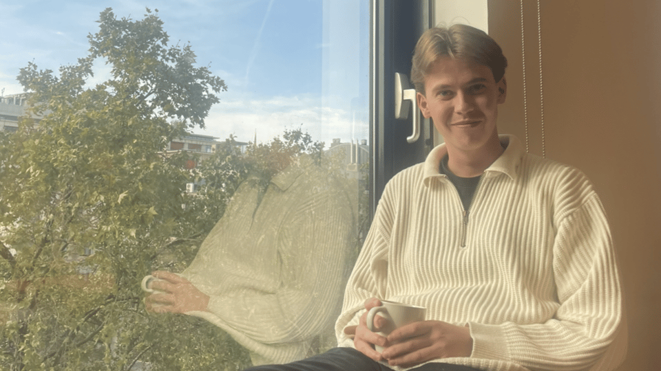 Ung man som sitter i ett fönster med en vit stickad tröja på sig och med en kaffekopp i handen. 