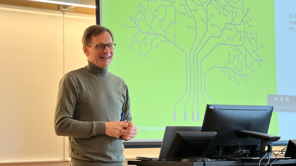 En man med grå tröja står och glasögon pratar framför en projektorduk