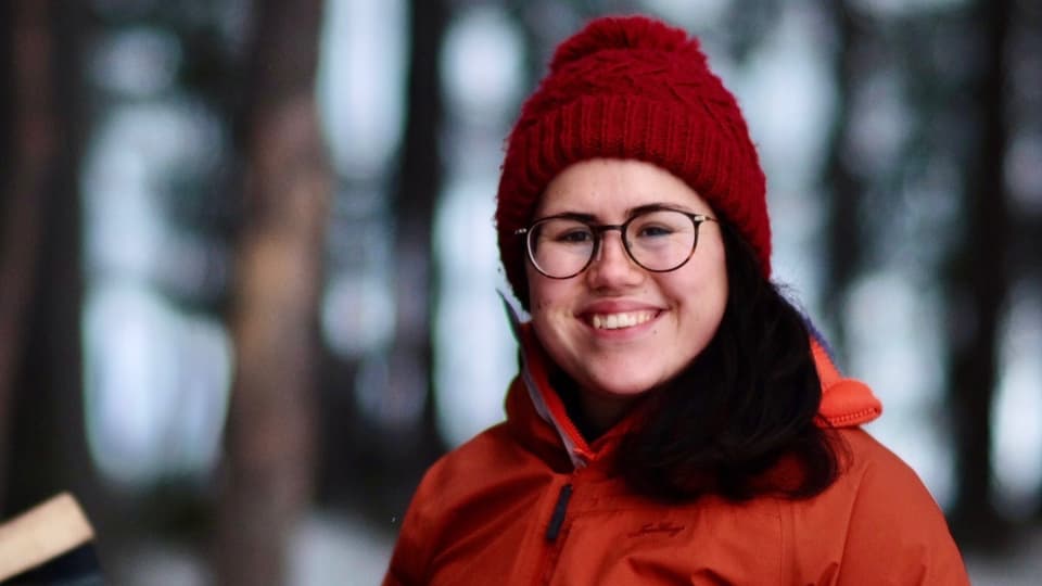 Kvinna med röd mössa och mörkbrunt hår iklädd en orange vinterjacka ler mot kameran. 