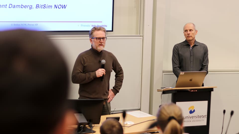 Två män står med mikrofon på scen. Publik i förgrunden. 