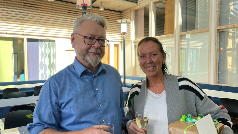 En mörkhårig kvinna i grå kofta står bredvid en gråhårig man i blå skjorta. Båda har champagneglas i händerna och ser glada ut. Kvinnan håller i ett paket.