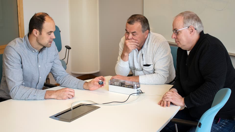 Three men sit at a table and discuss a prototype lying on the table.
