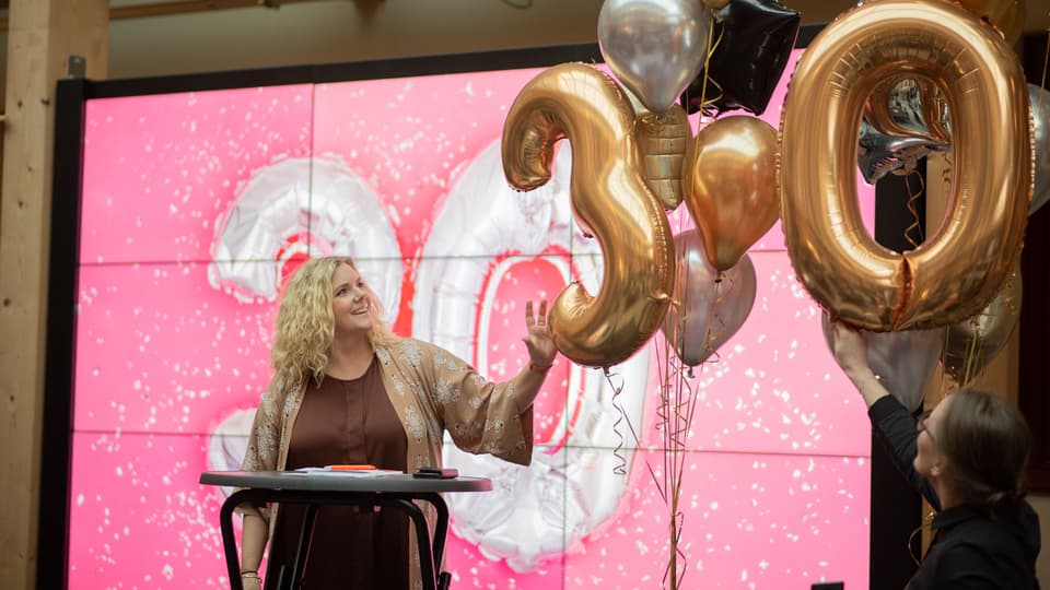 En kvinna står på en scen. Bredvid henne svävar ett antal ballonger i guld och silver. Två av ballongerna är formade som siffrorna 3 och 0. Hon tittar på en ballong och rör vid den.