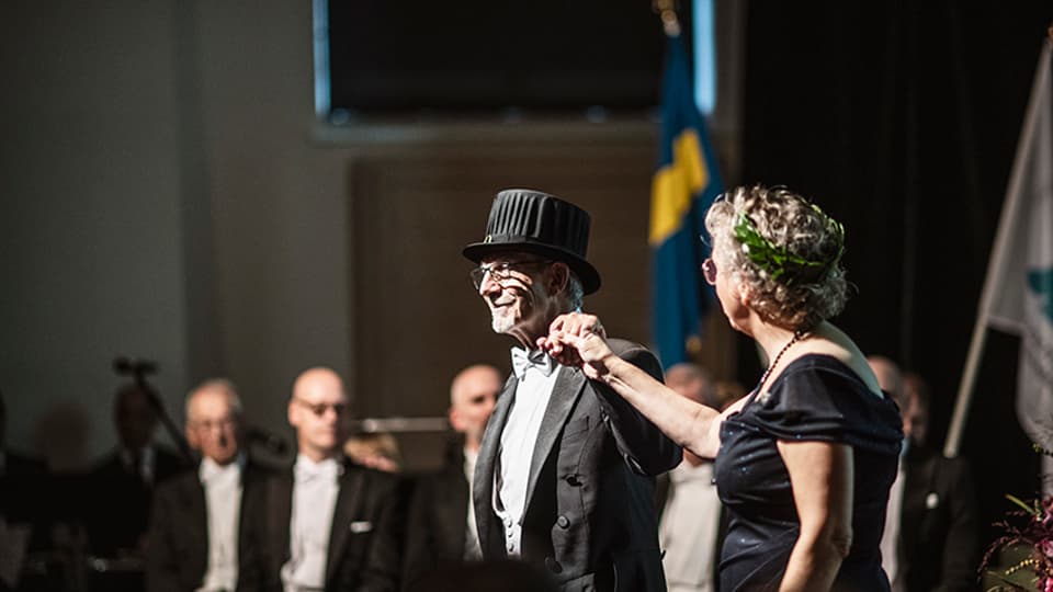 Två finklädda personer, en med doktorshatt och en med lagerkrans, står bredvid varandra på en scen och håller hand.