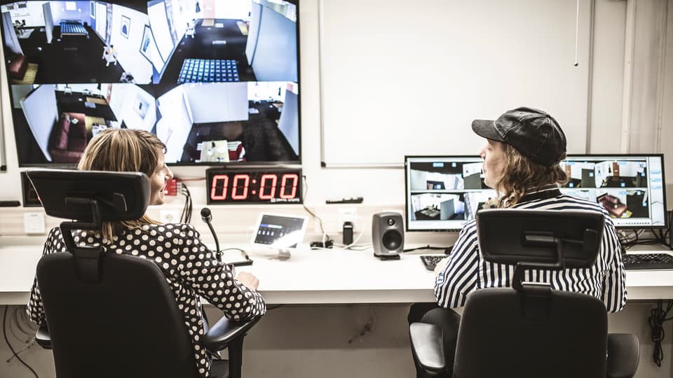 Två personer sitter vid ett långt arbetsbord. Framför dem finns flera datorskärmar, mikrofon och annan teknisk utrustning.