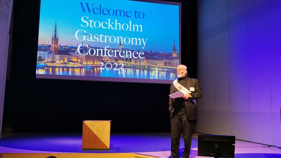 Man i kostym står på en scen med ett papper i handen och en skärm bakom sig med texten Stockholm Gastronomy Conference 2023