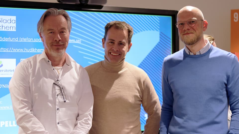 Three men stand in front of a large television screen.