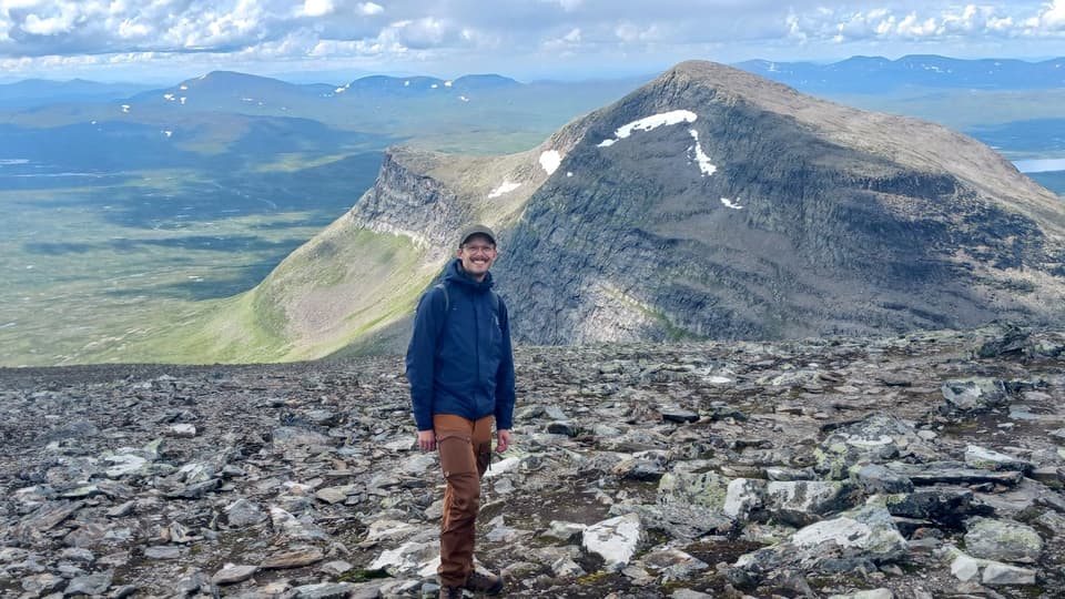 Man som i blå jacka, bruna byxor och keps står utomhus i stenigt landskap med berg i bakgrunden