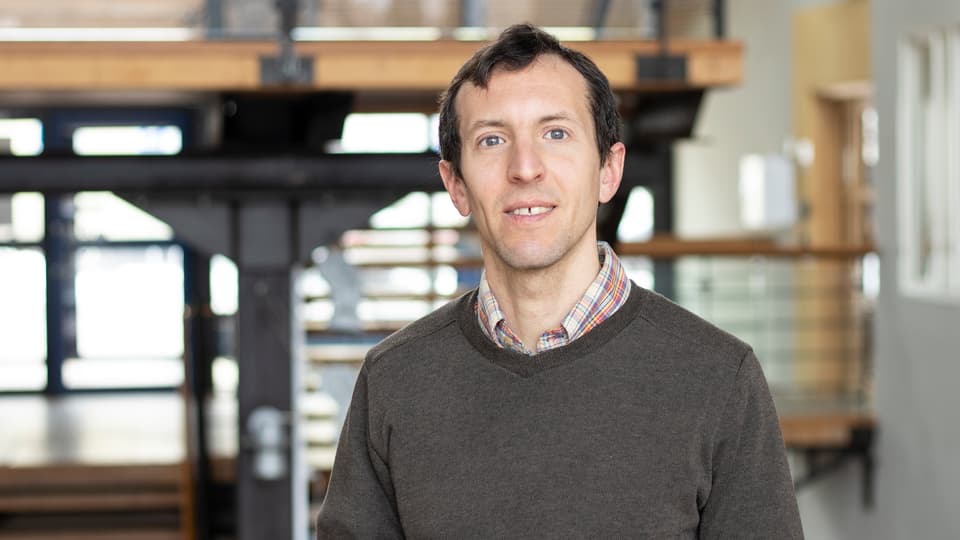 A man with dark hair, colorful shirt with a brown sweater over it.