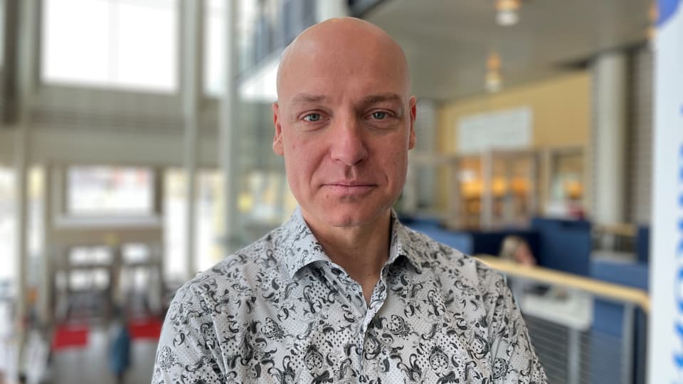 En man i mönstrad skjorta tittar in i kameran. Bakgrunden är suddig. 