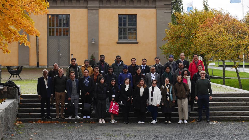 En grupp människor står på en stentrappa utomhus. I bakgrunden syns ett gult stenhus och träd med höstlöv.