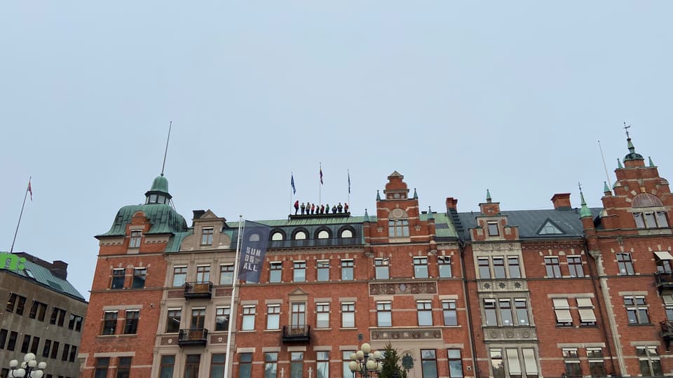 En grupp människor står på ett hustak. Huset är en pampig tegelbyggnad med valv och torn.