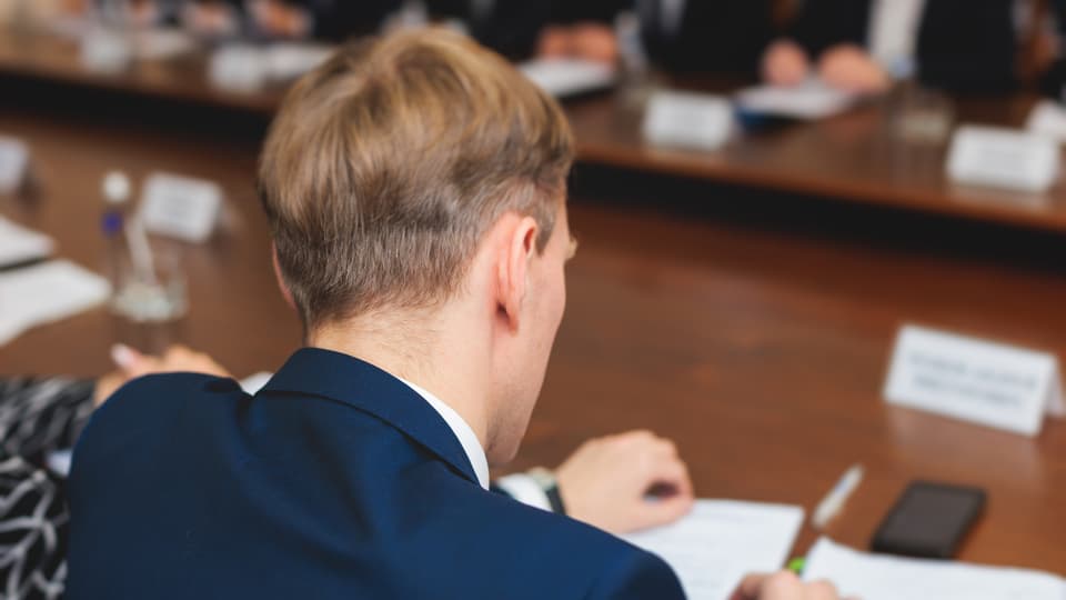 Man ser ryggen på en blond man i kavaj som sitter vid ett bord och skriver