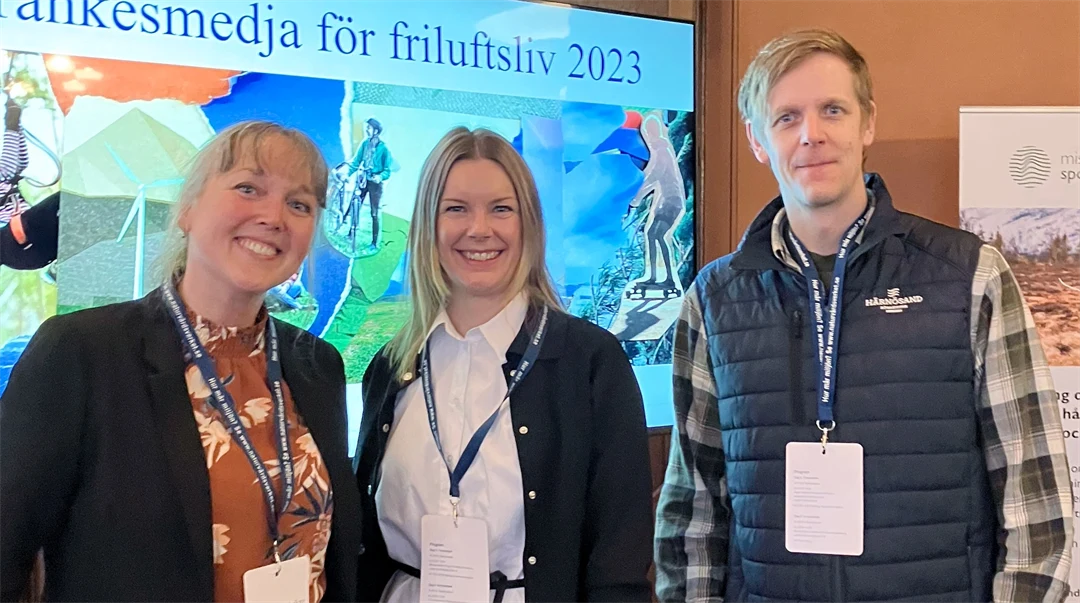 Två kvinnor och en man står i ett rum och tittar in i kameran. Bakom dem finns en skärm med en presentation på och de har namnskyltar hängande runt halsen 