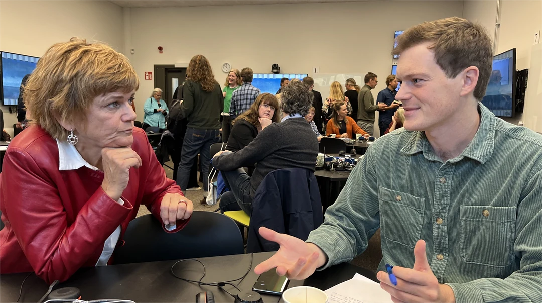 En kvinna och en man sitter vid ett bord och pratar med varandra. Kvinnan lyssnar och mannen ler och gestikulerar med båda händerna. 