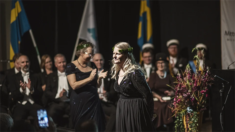 En finklädd kvinna med lagerkrans står på en scen. 