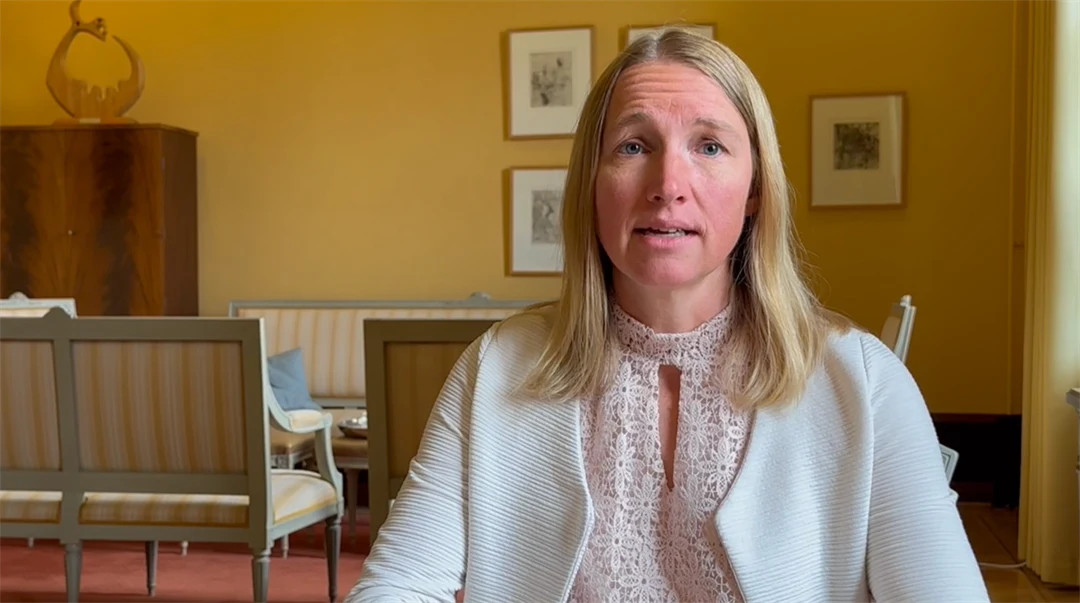 Blond kvinna med vit jacka sitter i ett rum med gula väggar och en sittgrupp i bakgrunden. Tavlor hänger på väggarna. 