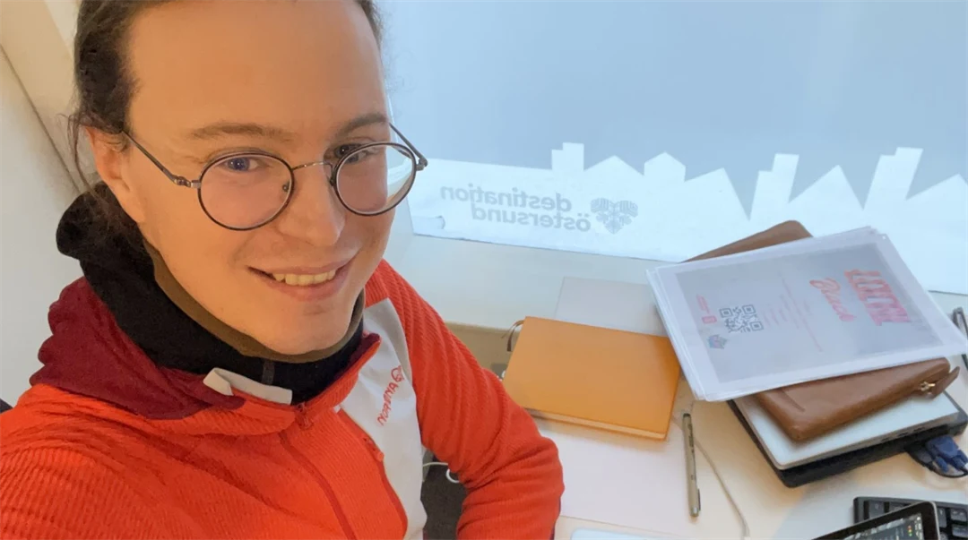 Ung man i glasögon och orange tröja som sitter med sin dator vid ett skrivbord  