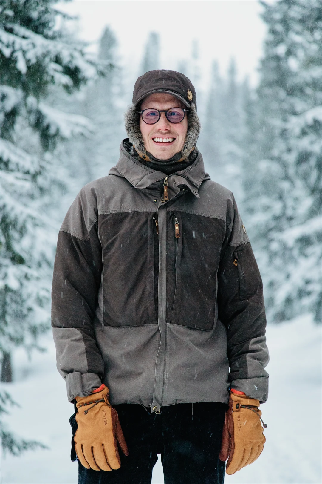 Happy young man in thick winter clothes standing outdoors in a winter landscape with snow-covered fir trees in the background.