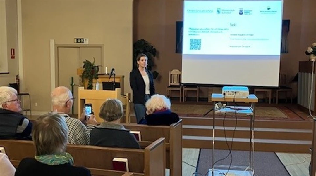 En kvinna står längst fram i en lokal med en en digital skärm. Människor sitter i bänkar i publiken och lyssnar.