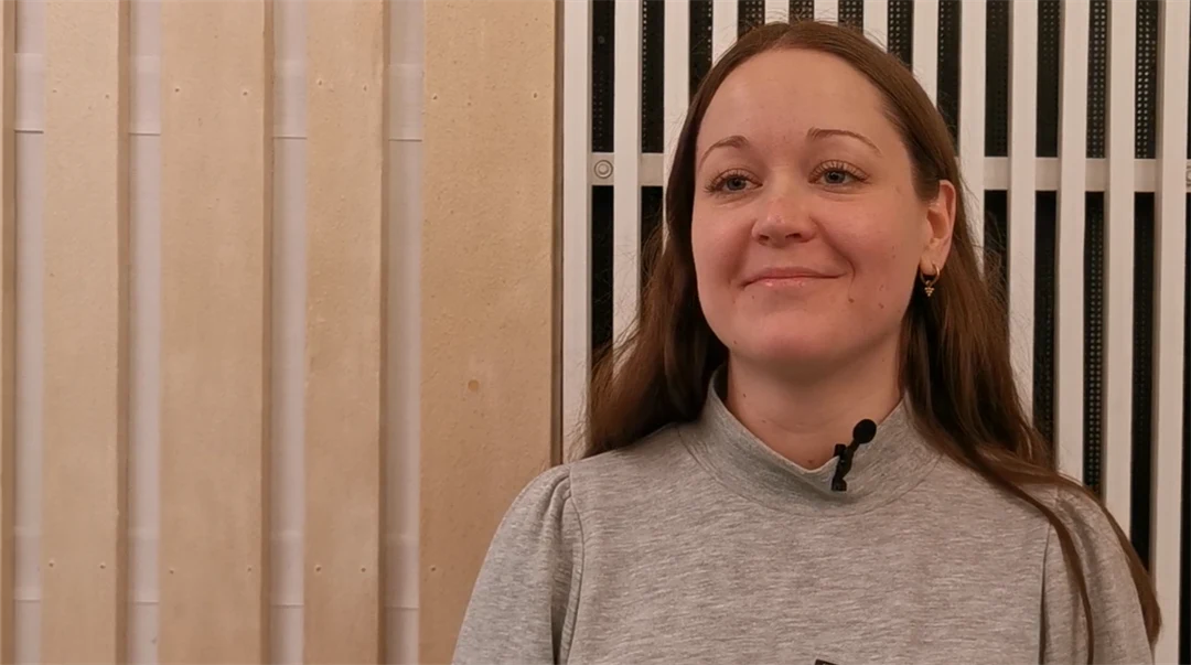 Kvinna med långt brunt hår och en grå tröja står framför en vägg med träpanel