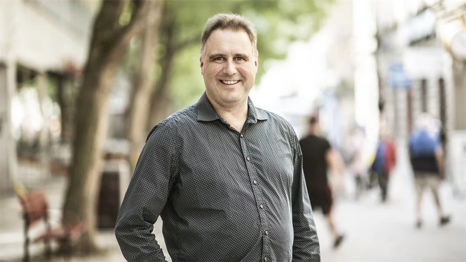 Man med ljusbrunt hår, klädd i en grå skjorta ler mot kameran