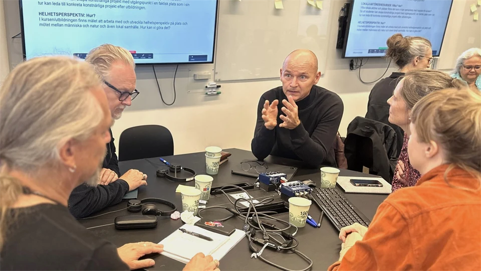 Fem personer sitter runt ett bord och en av dem, en man med svart tröja gestikulerar med händerna medan han pratar