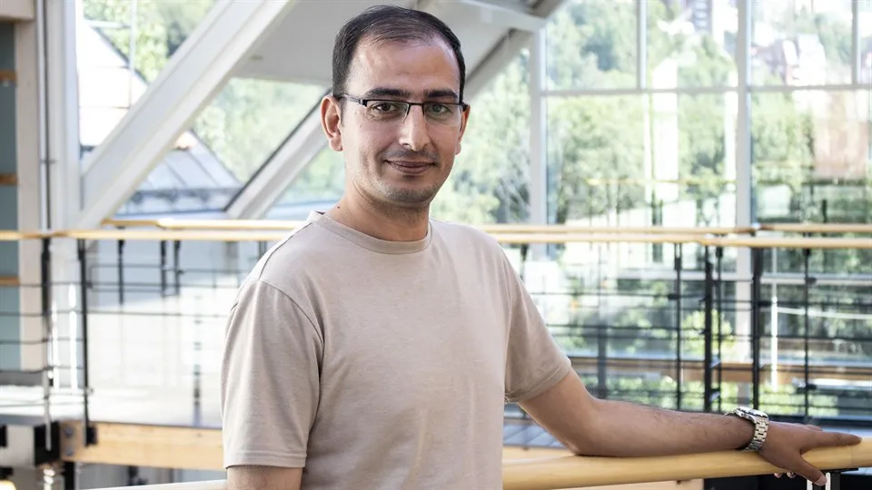 En man i glasögon, mörkt hår och beige t-shirt poserar framför kameran.