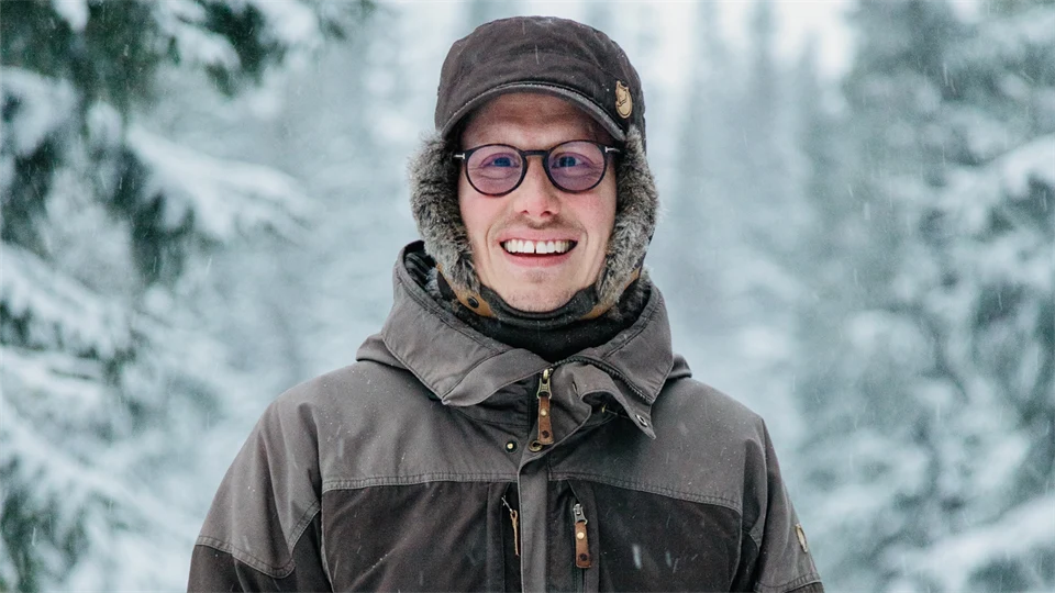 Glad ung man i tjocka vinterkläder står utomhus i vinterlandskap med snöklädda granar i bakgrunden