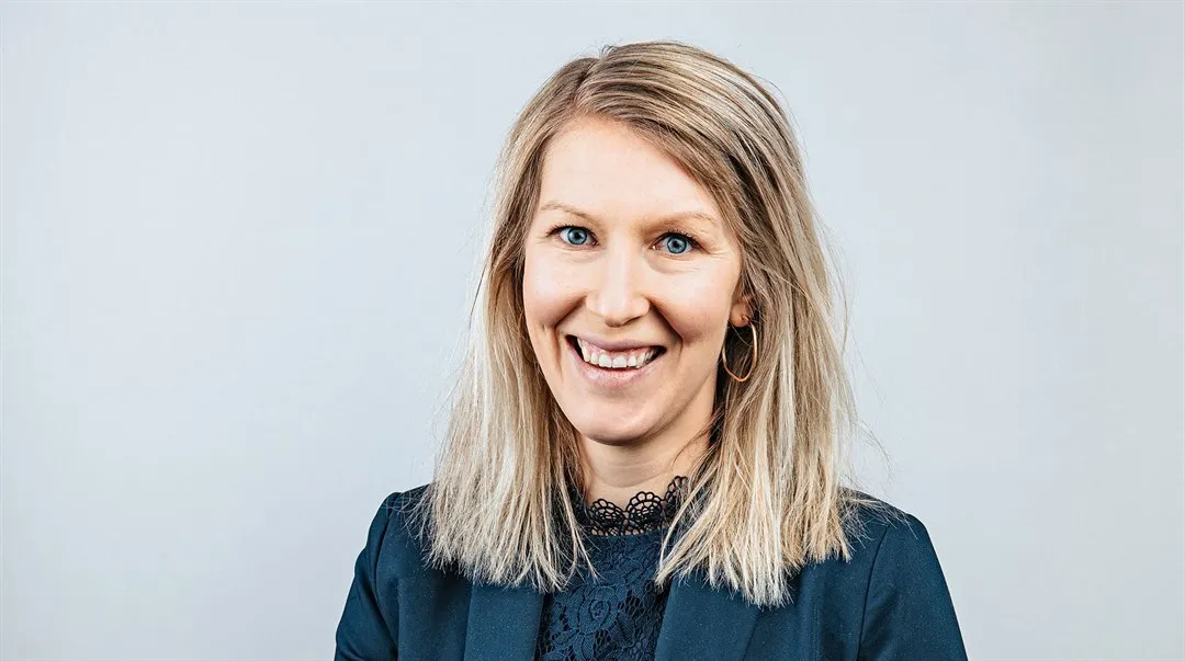 En kvinna i blå kavaj och med ett stort leende som sitter på en pall med händerna i knät