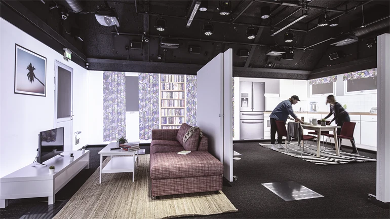 Ett stort rum med svart golv och tak. Rummet ser ut som en lägenhet med mellanväggar, möbler, och projiceringar på väggarna. Två personer står vid köksbordet. I taket finns massor av teknisk utrustning.