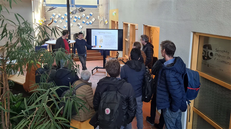 En grupp människor som står med ryggen mot kameran och tittar på en tv och en man som presenterar. 