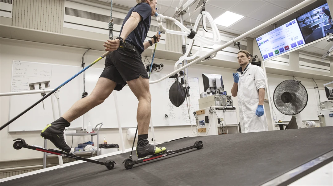 Roller skier with hoses from a face mask on a treadmill supervised by test leader.