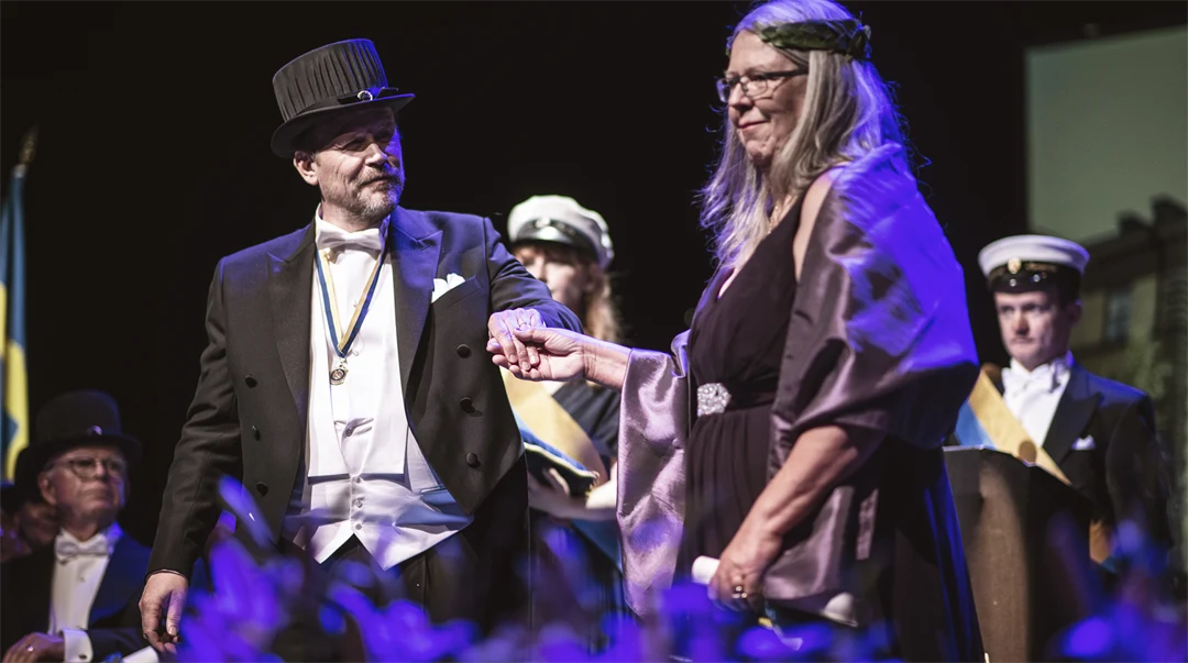 Two well-dressed people - one with a doctor's hat, one with a laurel wreath holding hands.