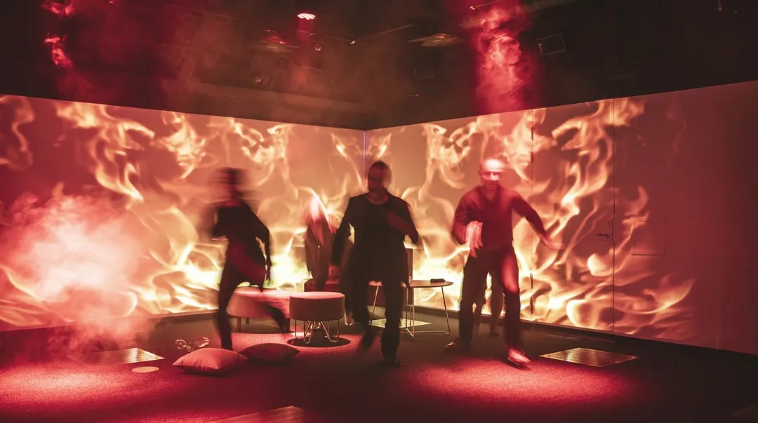 A large room with flames projected on the walls. The light is dimmed and red-toned. Four people are running.