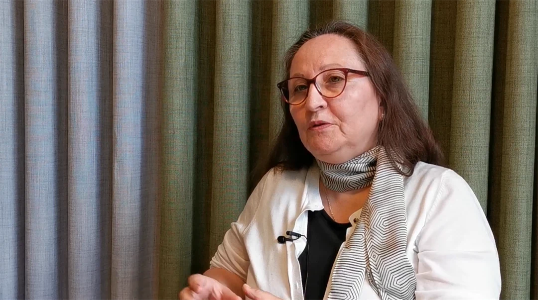 Kvinna med brunt långt hår klädd i vit blus och grön scarf blir intervjuad