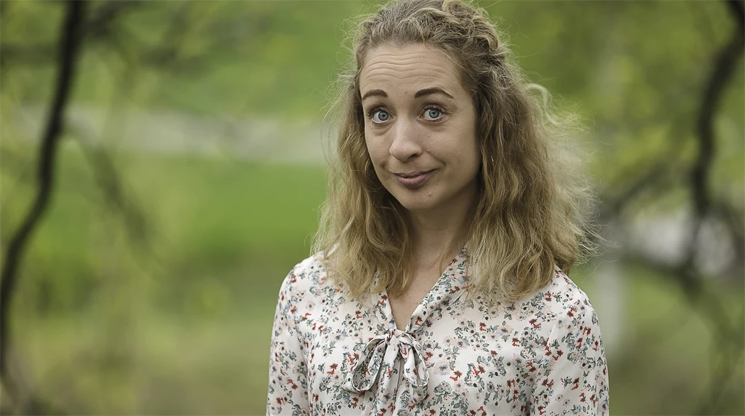 Kvinna med lockigt axellågt hår och blommig blus står utomhus med grönskande träd runt om kring sig.