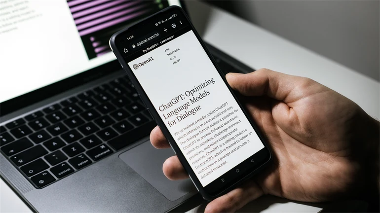 Hand som håller i en mobiltelefon med texten ChatGPT en dator i bakgrunden. 