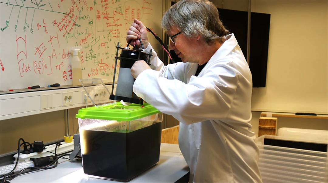 A man in a lab coat lowers an instrument into a small aquarium filled with dark liquid.