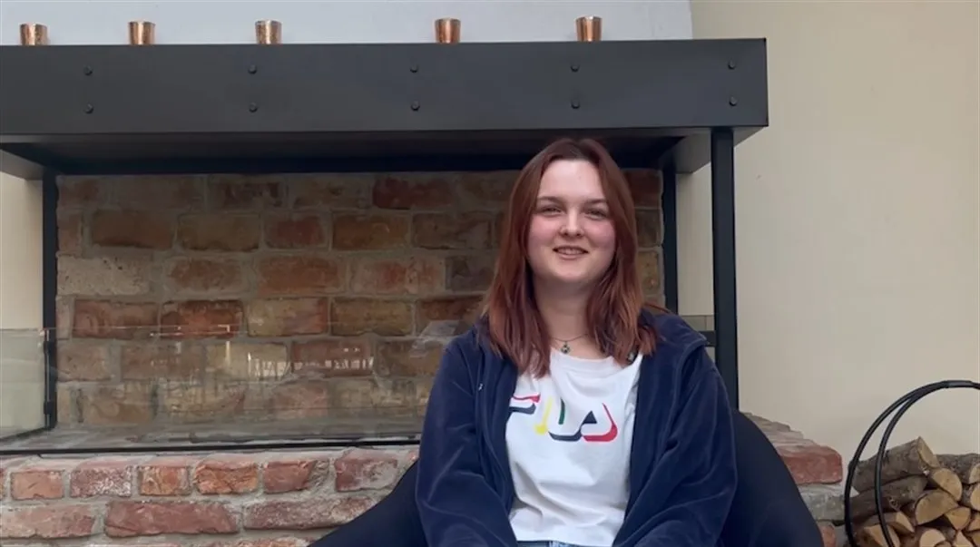 Kvinna sitter framför en eldstad, hon ler och tittar in i kameran