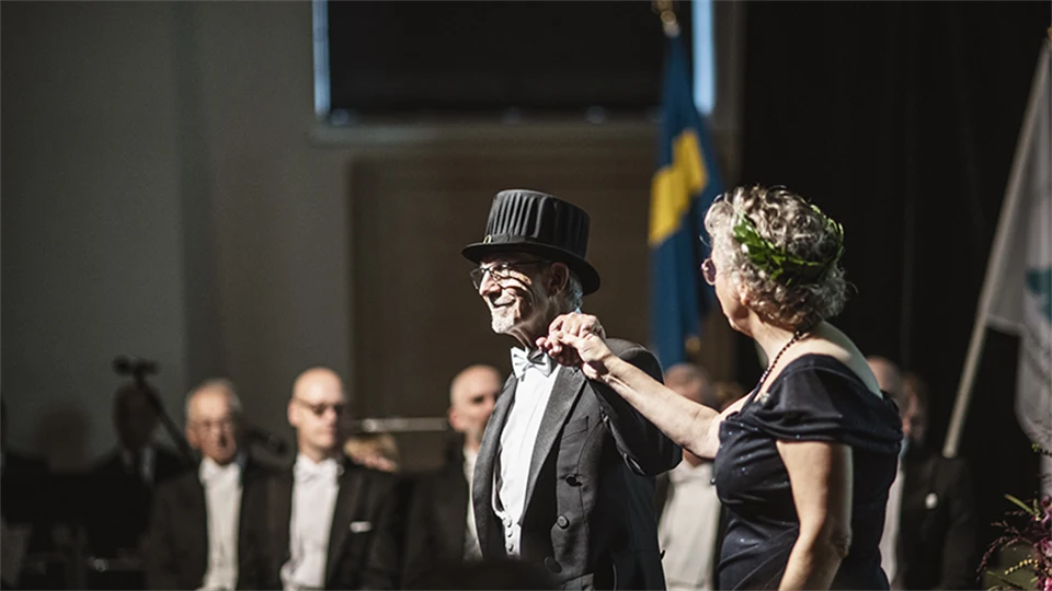 Två finklädda personer, en med doktorshatt och en med lagerkrans, står bredvid varandra på en scen och håller hand.