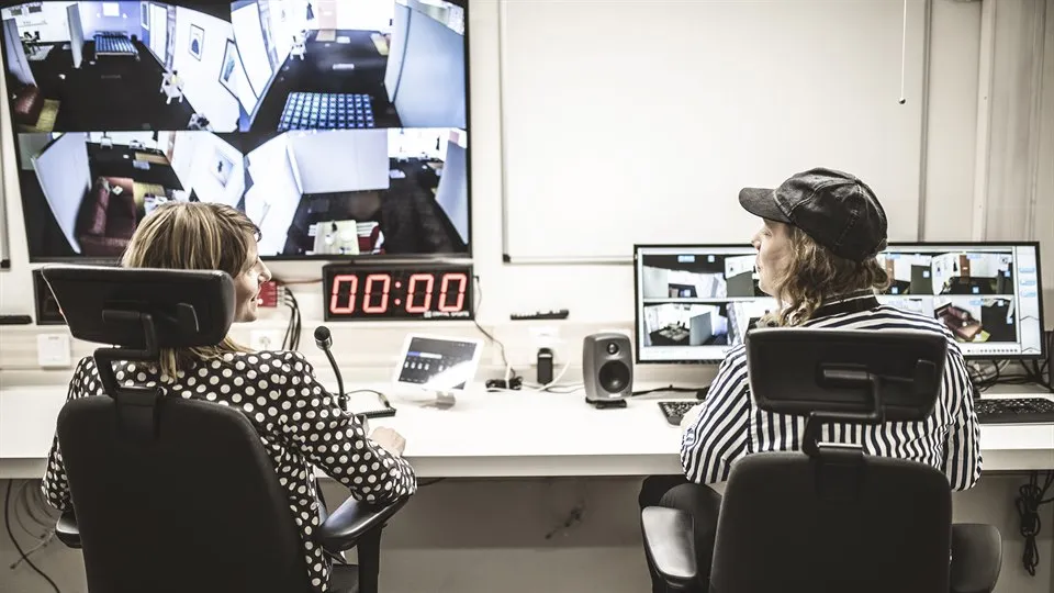 Två personer sitter vid ett långt arbetsbord. Framför dem finns flera datorskärmar, mikrofon och annan teknisk utrustning.