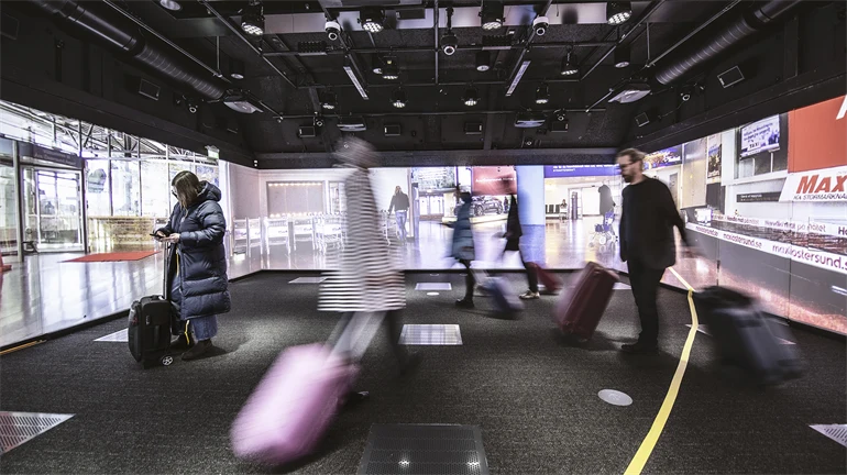 Flera personer står i ett rum med bilder från en flygplats projicerade på väggarna. De har ytterkläder och resväskor. På golvet sitter en gul tejpremsa. I taket finns massor av teknisk utrustning.