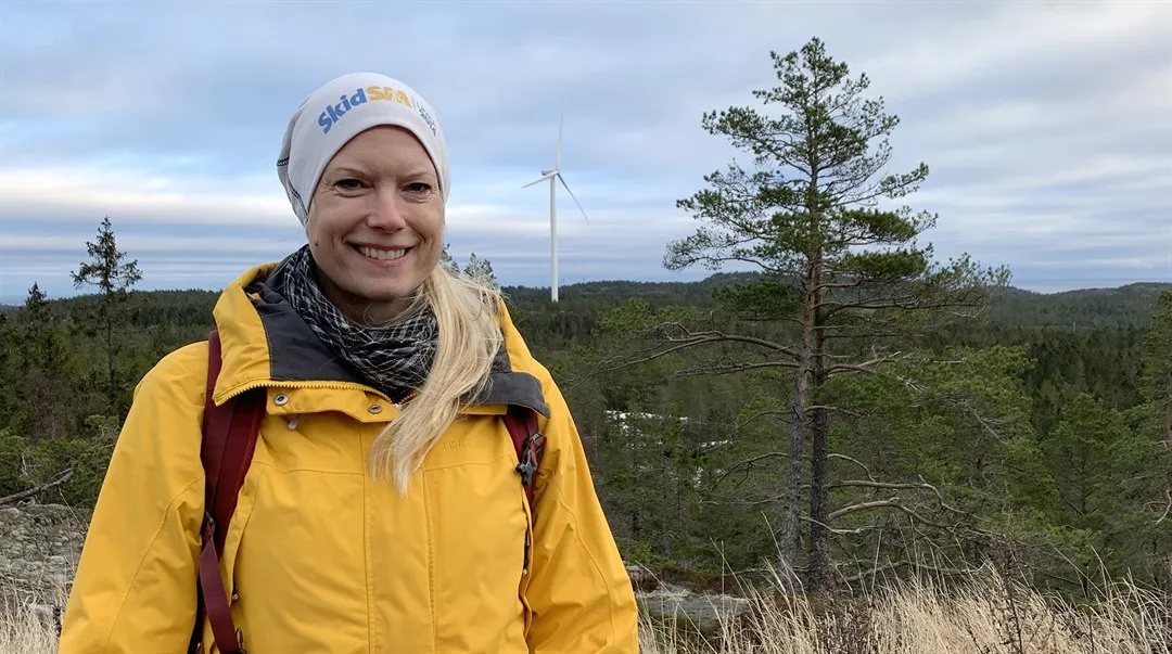 Kvinna i gul jacka och vit mössa stående framför träd och ett vindkraftverk.